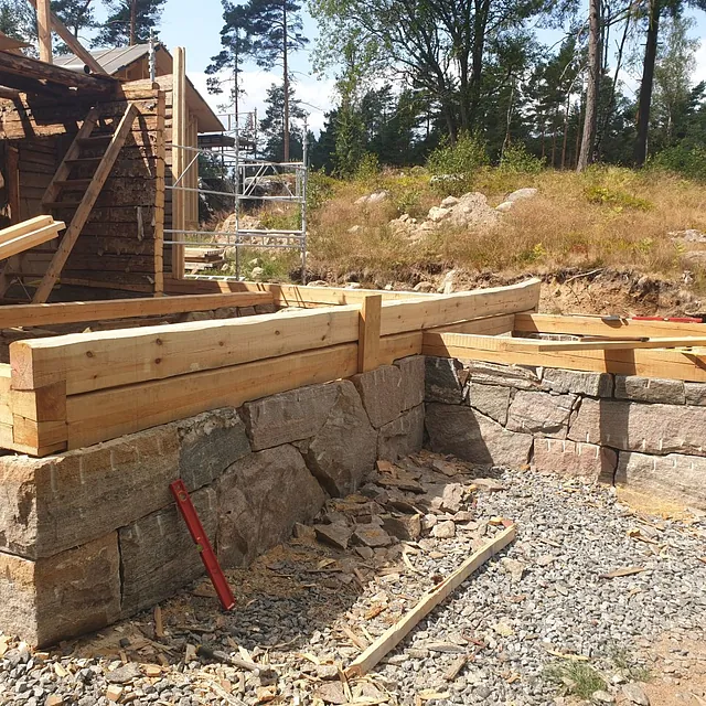 Pågående byggnation med träkonstruktion, stenmur och arbetsmaterial.